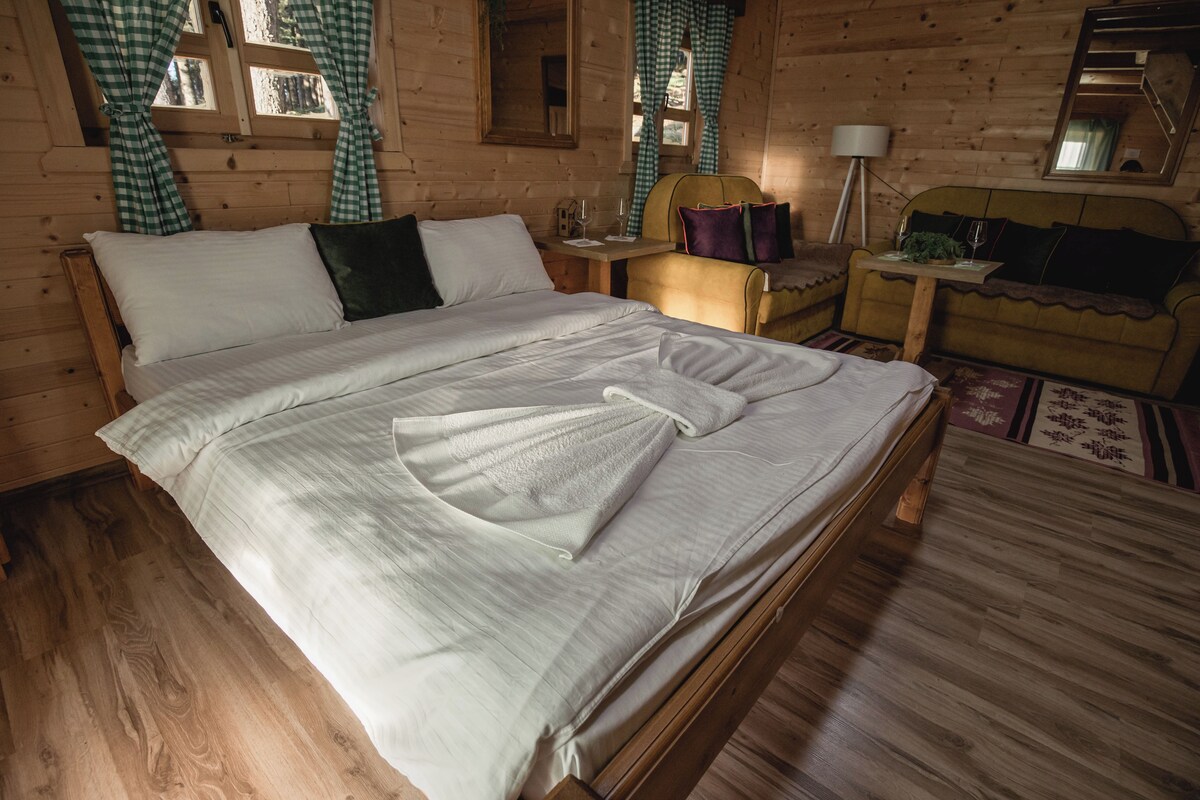 A wooden cabin bedroom with a large bed, a nightstand, a chair, and a small table with a lamp.