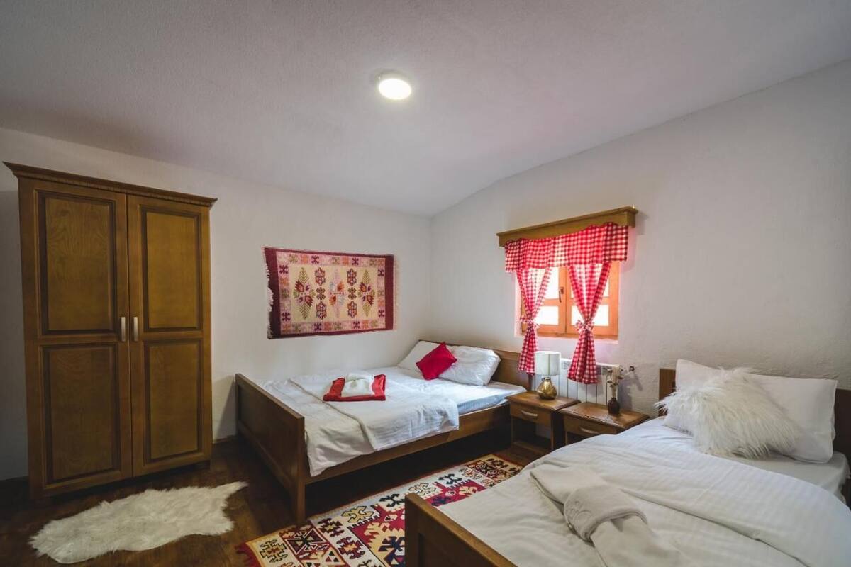 A bedroom with two beds, a wardrobe, a patterned wall hanging, and a window with red checkered curtains.