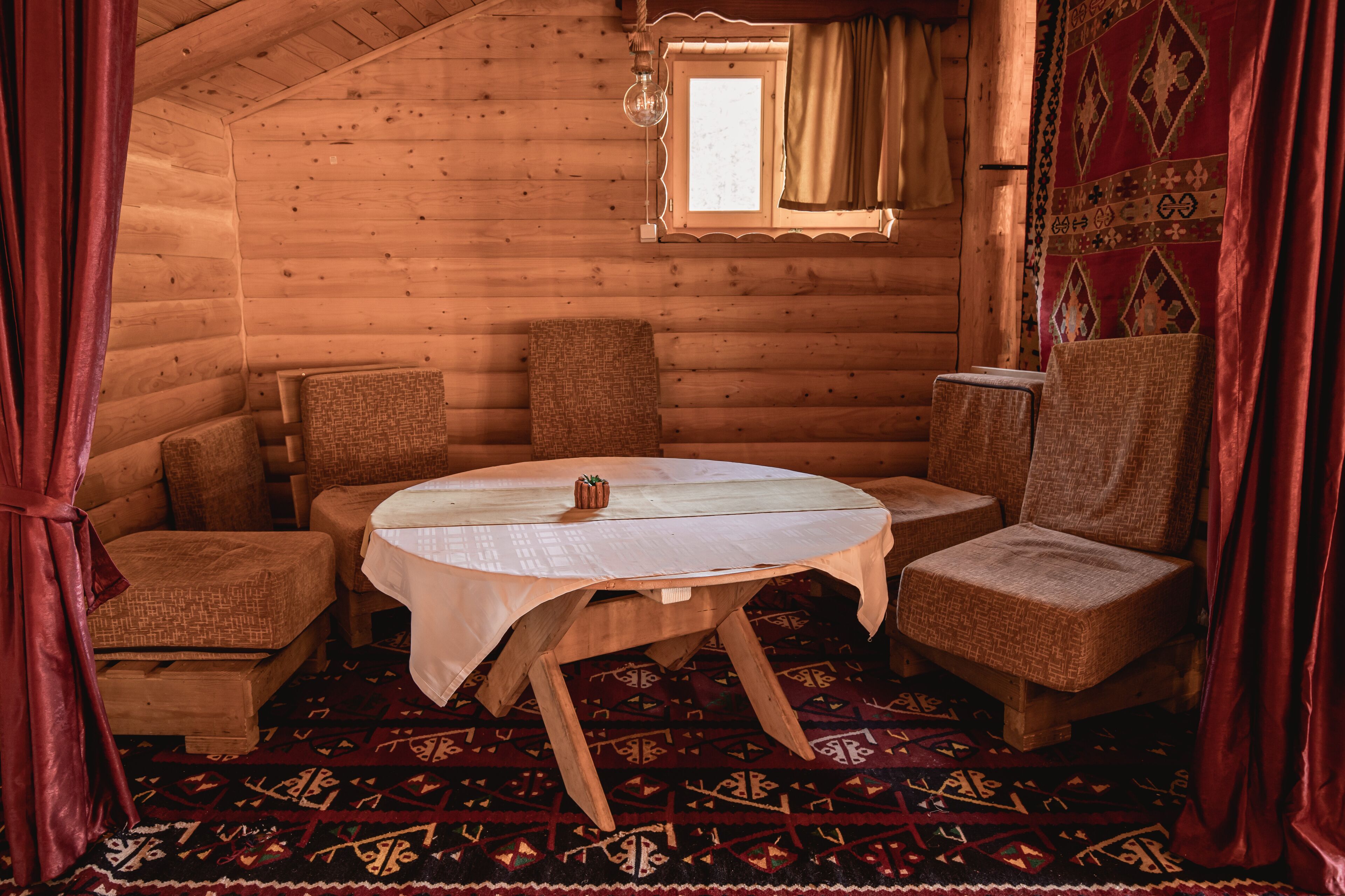 A cozy log cabin room with a round wooden table, four woven chairs, a patterned rug, and a window with curtains.