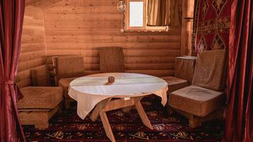 A cozy log cabin room with a round wooden table, four woven chairs, a patterned rug, and a window with curtains.