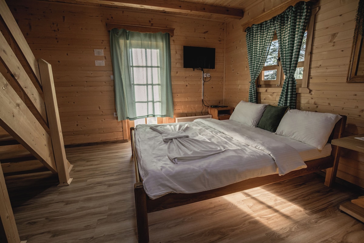 A wooden cabin bedroom with a bed, a television, a window with curtains, and a staircase.