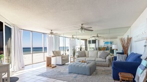 A spacious living room with a large window overlooking the beach, a ceiling fan, and a blue sofa.