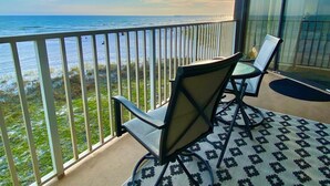A balcony with two chairs and a table, overlooking a beach and ocean.