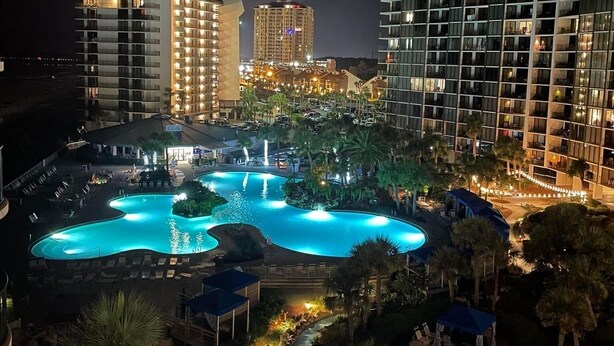 A nighttime view of a hotel with a large, illuminated swimming pool surrounded by palm trees and lounge chairs.