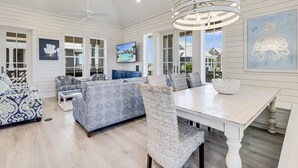 A spacious living room with a large sofa, a dining table, and wicker chairs. There is a TV mounted on the wall, and a ceiling fan is visible.