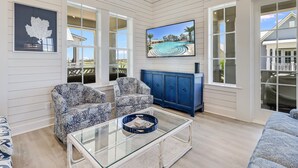 A spacious living room with a glass coffee table, patterned armchairs, a blue cabinet, and a large TV displaying a pool scene.