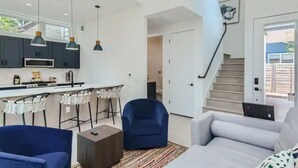 Une cuisine moderne avec des meubles de couleur sombre, un plan de travail blanc et des luminaires suspendus. On y trouve un coin repas avec des tabourets de bar et une chaise bleue. Un escalier mène à l’étage supérieur.