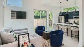 Un salon moderne avec un canapé, une table basse et un téléviseur fixé au mur. On y trouve également un coin cuisine avec des tabourets de bar et une grande fenêtre.
