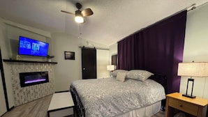 Une chambre à coucher comprenant un lit, un téléviseur, un ventilateur de plafond, un radiateur fixé au mur, une table de chevet en bois et une fenêtre avec des rideaux violets.