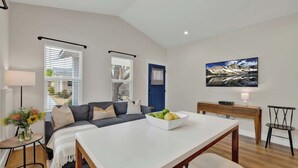 Una sala de estar moderna con un sofá, una mesa de comedor con un cuenco de fruta, un televisor en la pared y una ventana con persianas.