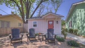 Una piccola casa rosa con una porta rossa, due poltrone Adirondack e un tavolo di fronte.