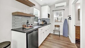 Una cucina compatta con lavastoviglie, mobili bianchi, una mensola in legno e una finestra con persiane.