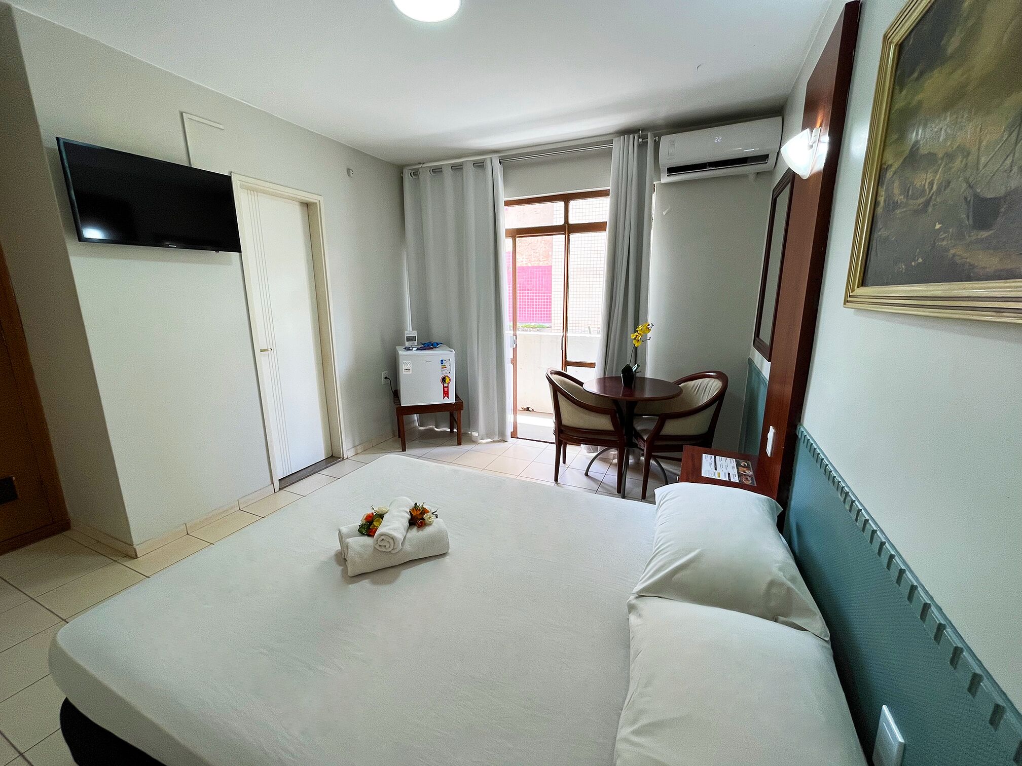 A hotel room with a bed, a desk, a chair, a TV, and an air conditioning unit.