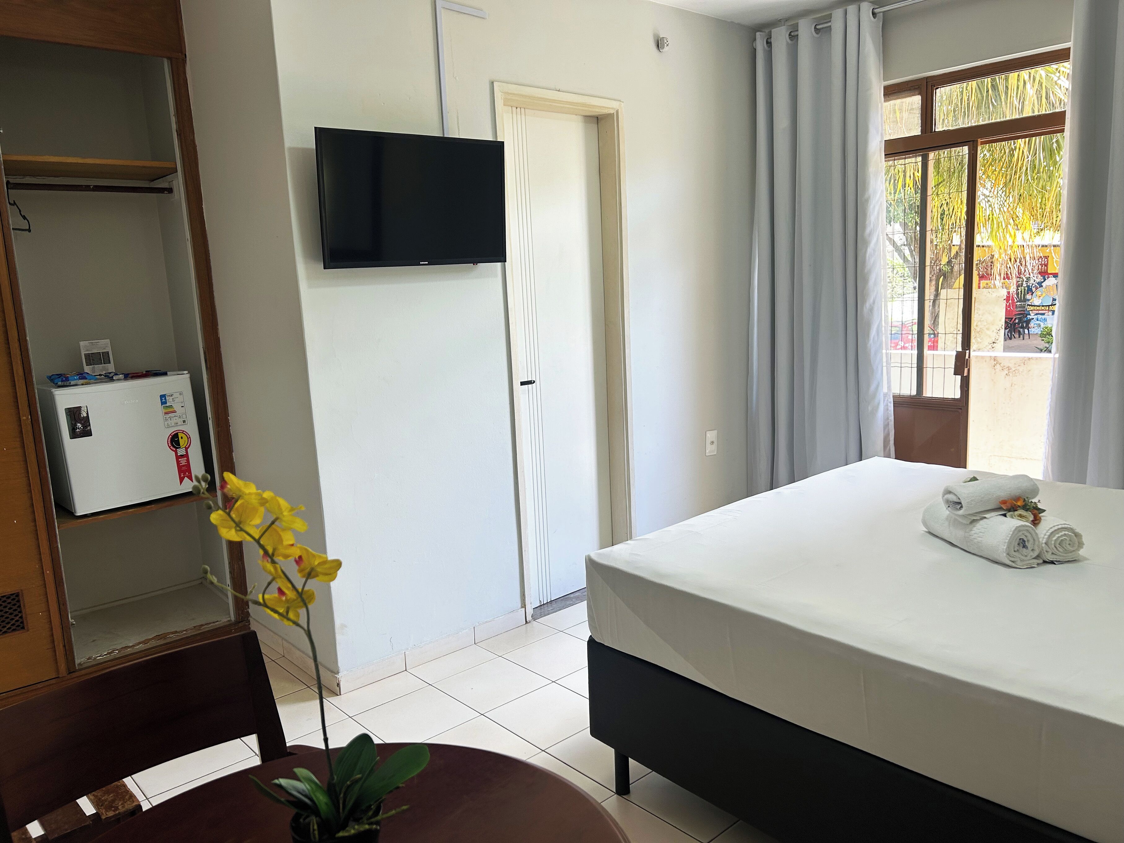 A hotel room with a bed, a television mounted on the wall, a small refrigerator, and a window with curtains.
