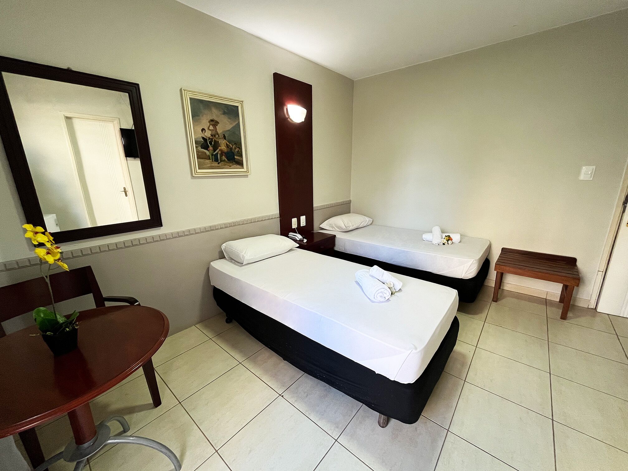 A hotel room with two beds, a desk, a chair, a small table, a mirror, and a painting on the wall.