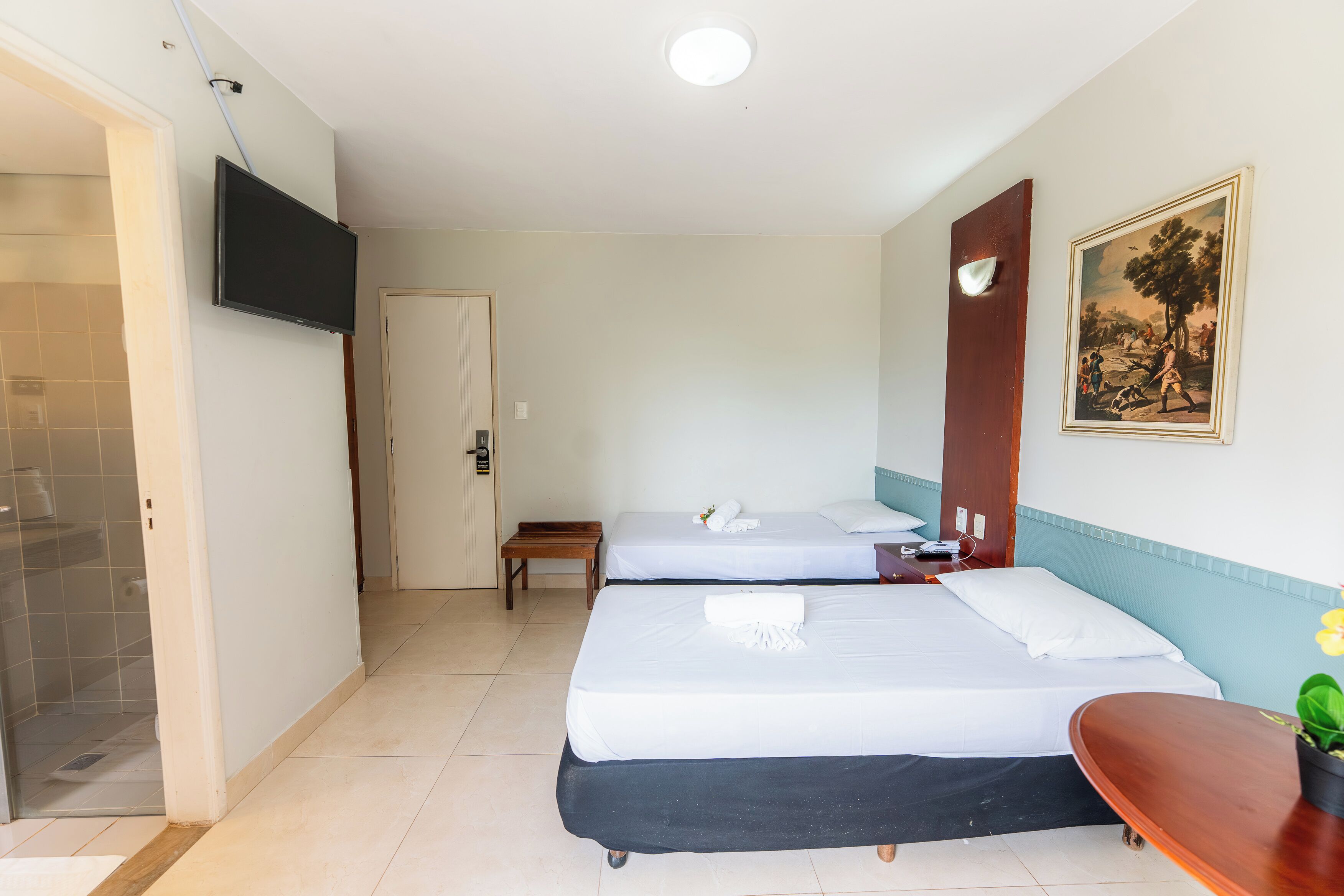 A hotel room with two beds, a TV, a painting, and a small table with a plant.