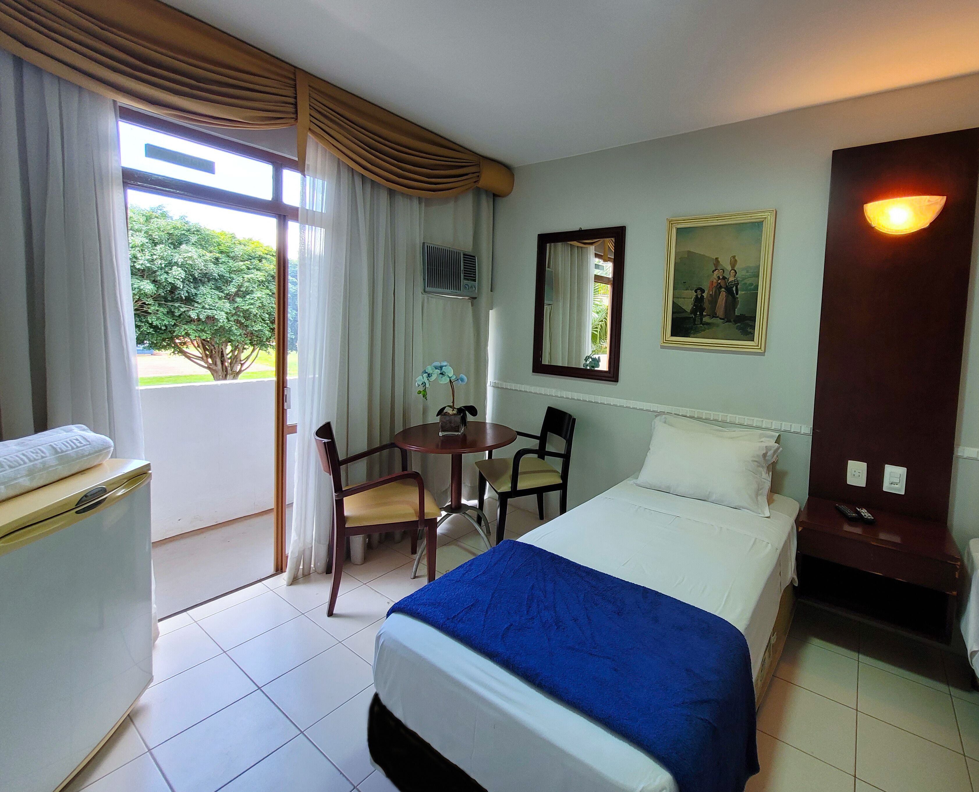 A hotel room with a bed, a desk with a chair, a window with curtains, a painting on the wall, and a nightstand with a lamp.