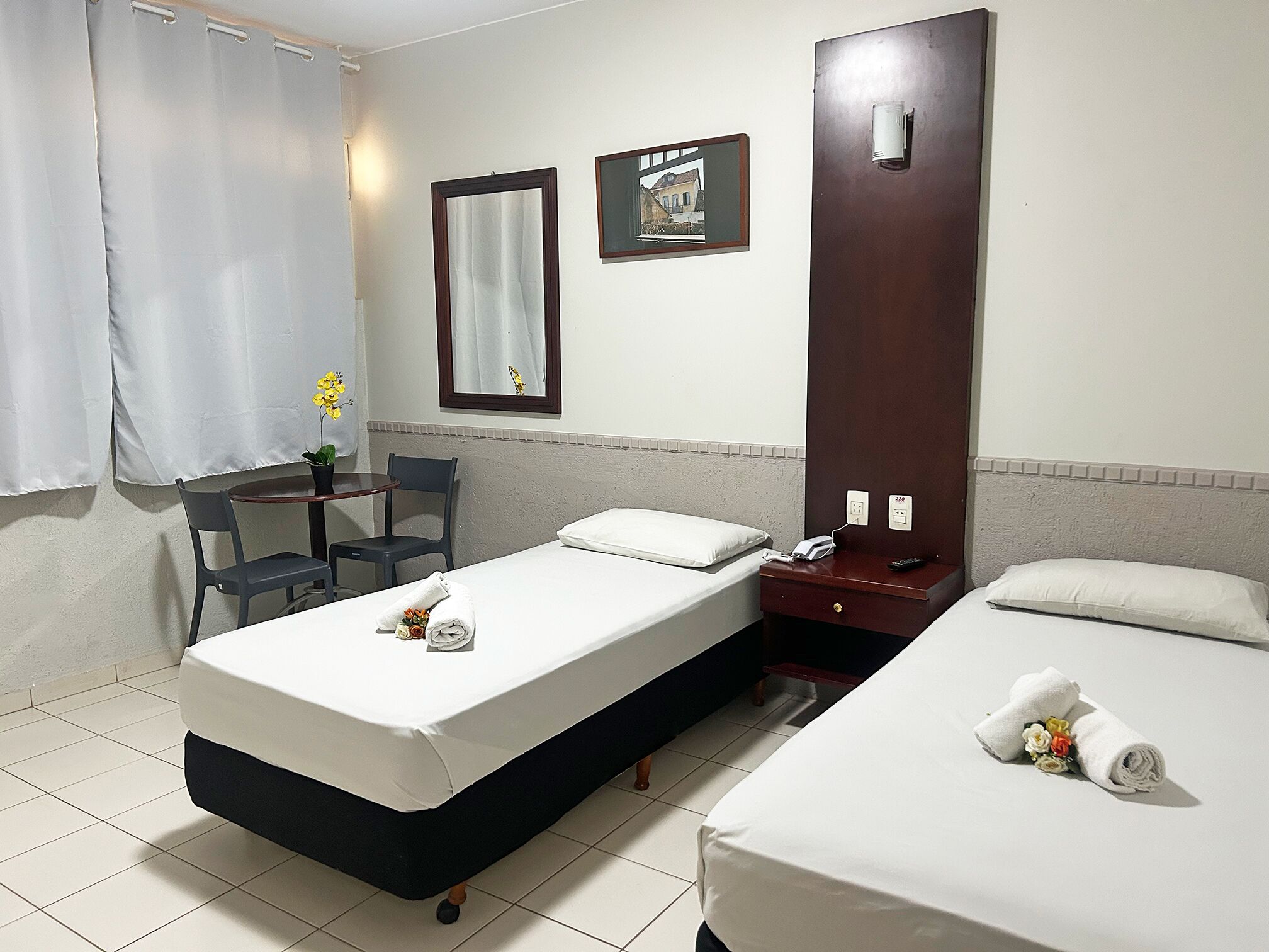 A hotel room with two beds, a desk, a chair, a small table with a potted plant, a mirror, and a picture on the wall.