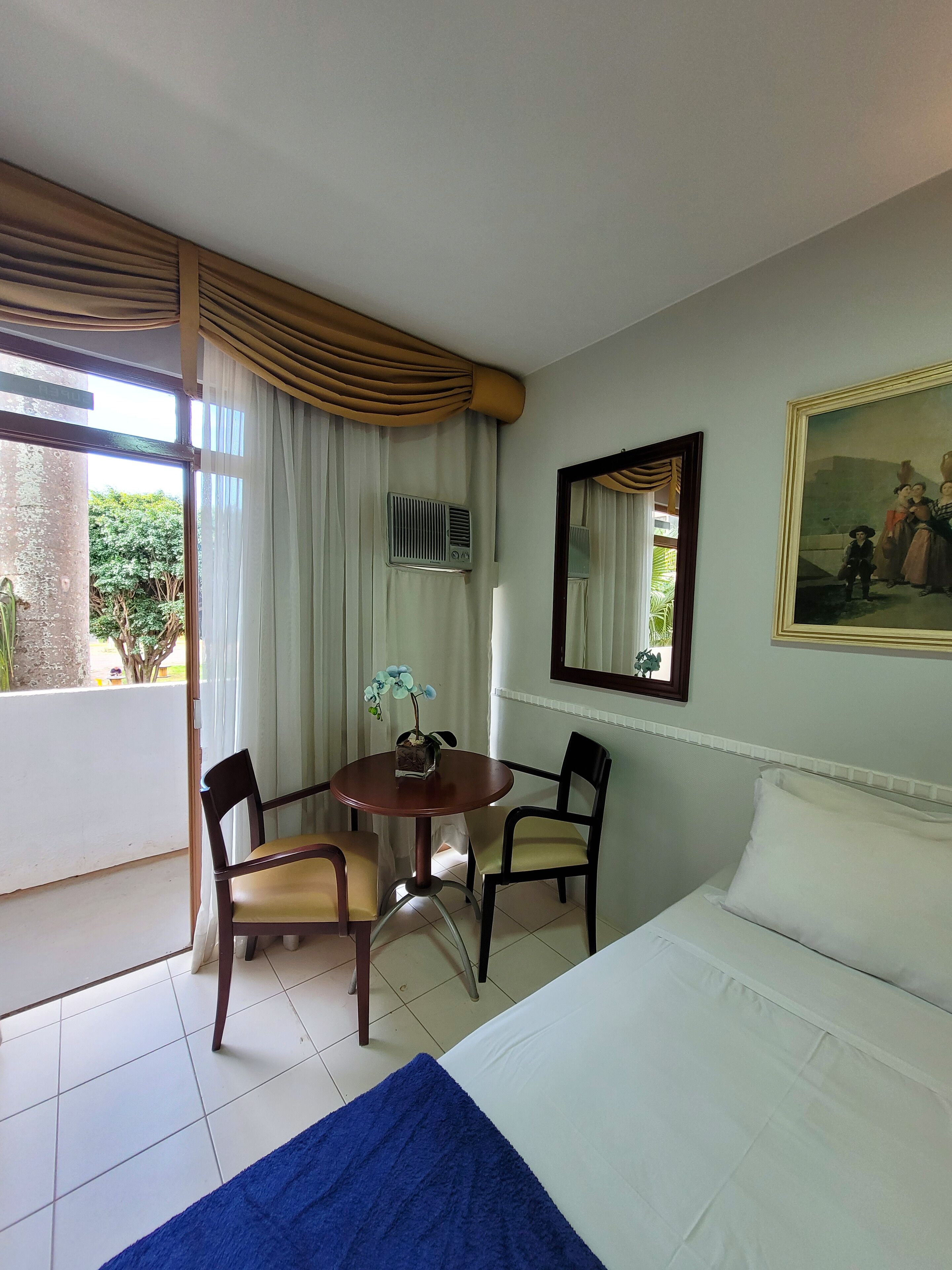 Habitación de hotel con cama, una mesa de comedor con sillas, ventana con cortinas, aire acondicionado montado en la pared y un cuadro.