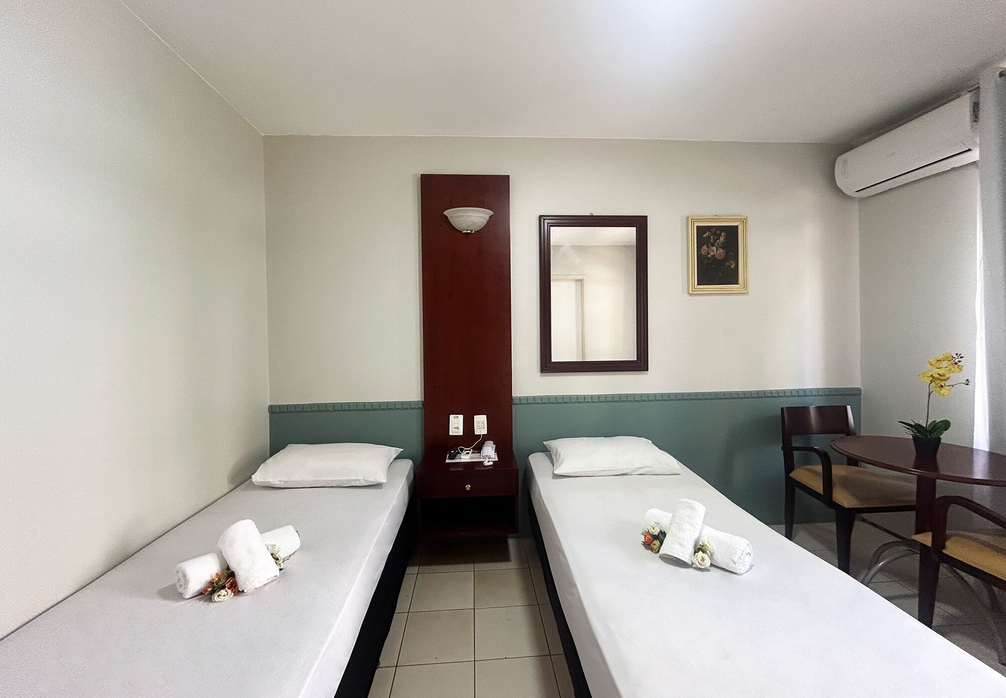 A hotel room with two single beds, a small table, a chair, a desk, a mirror, and an air conditioning unit.