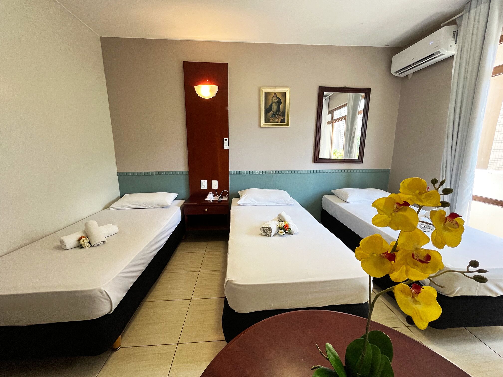 A hotel room with two single beds, a small table, a potted orchid, a painting, and an air conditioning unit.