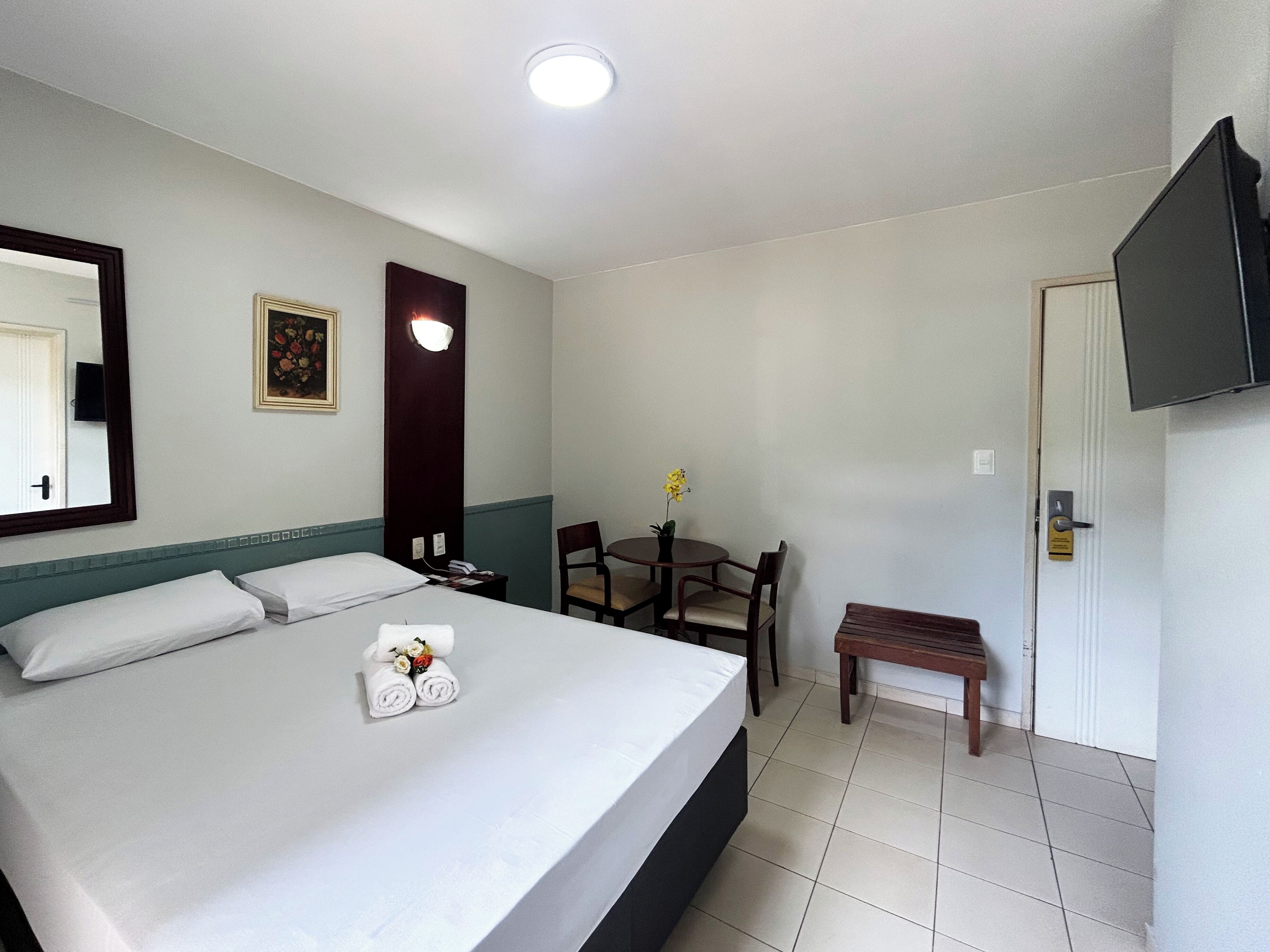 Quarto de hotel com cama, escrivaninha, cadeira, mesa pequena, televisão e porta.