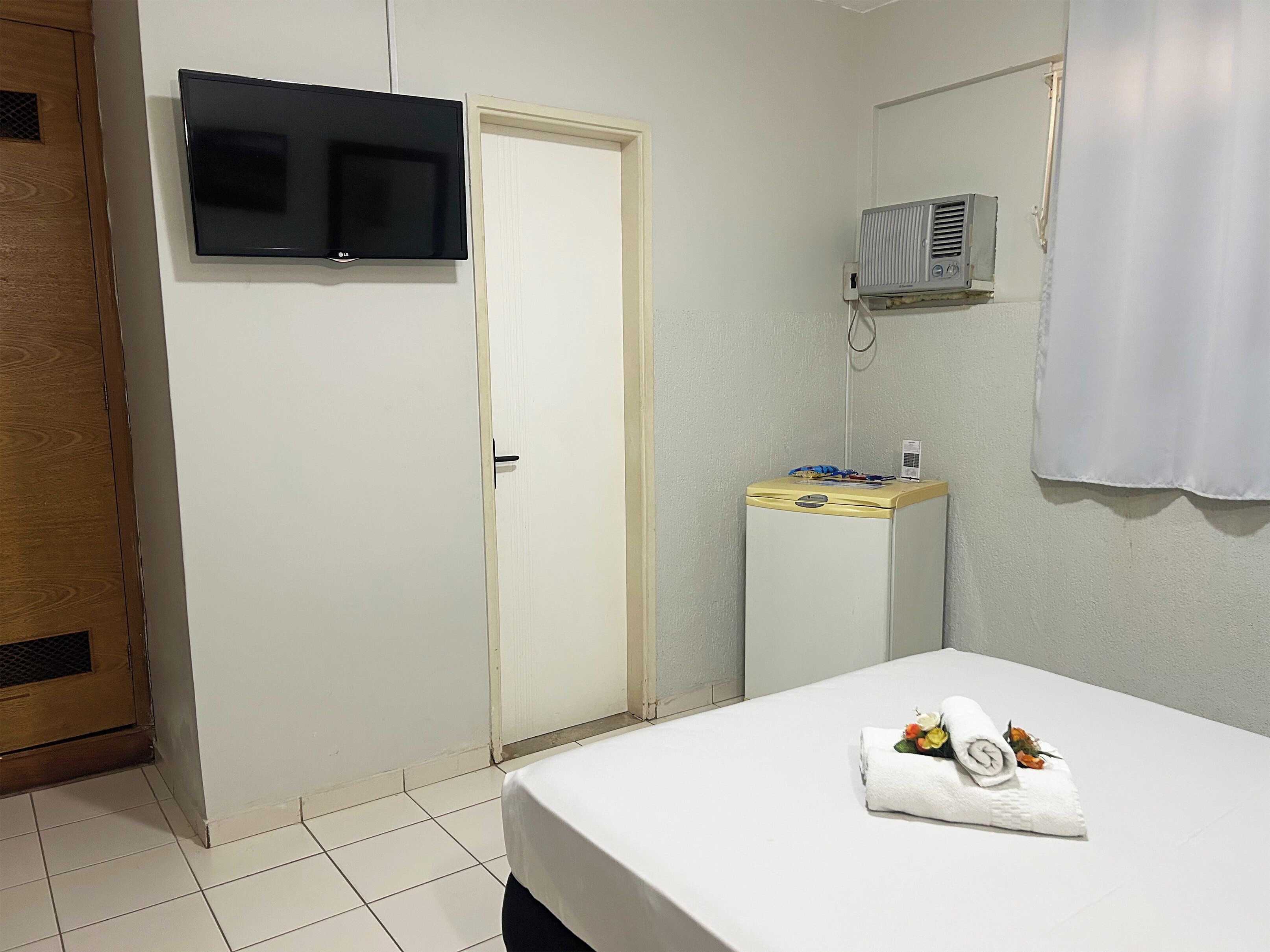 Una habitación de hotel con cama, televisor montado en la pared, equipo de aire acondicionado y armario de madera.