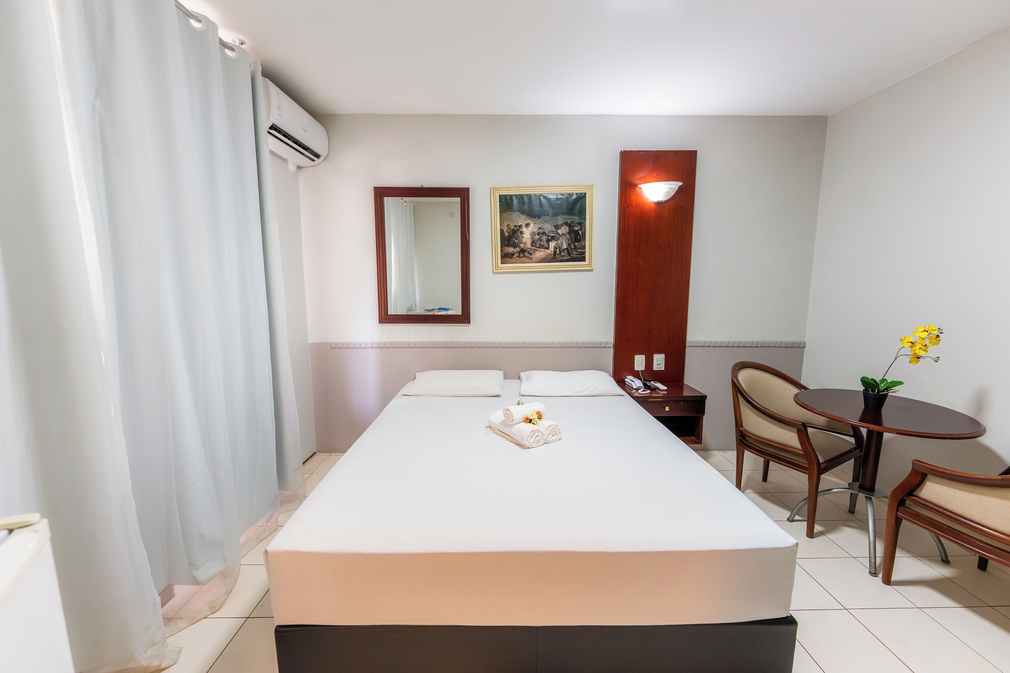 A hotel room with a bed, a bedside table, a chair, a small table with a plant, a mirror, and a picture on the wall.