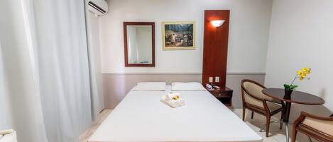 A hotel room with a bed, a bedside table, a chair, a small table with a plant, a mirror, and a picture on the wall.