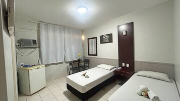 A hotel room with a bed, a desk, a chair, a small table with a flower, a mirror, and a wall-mounted heater.