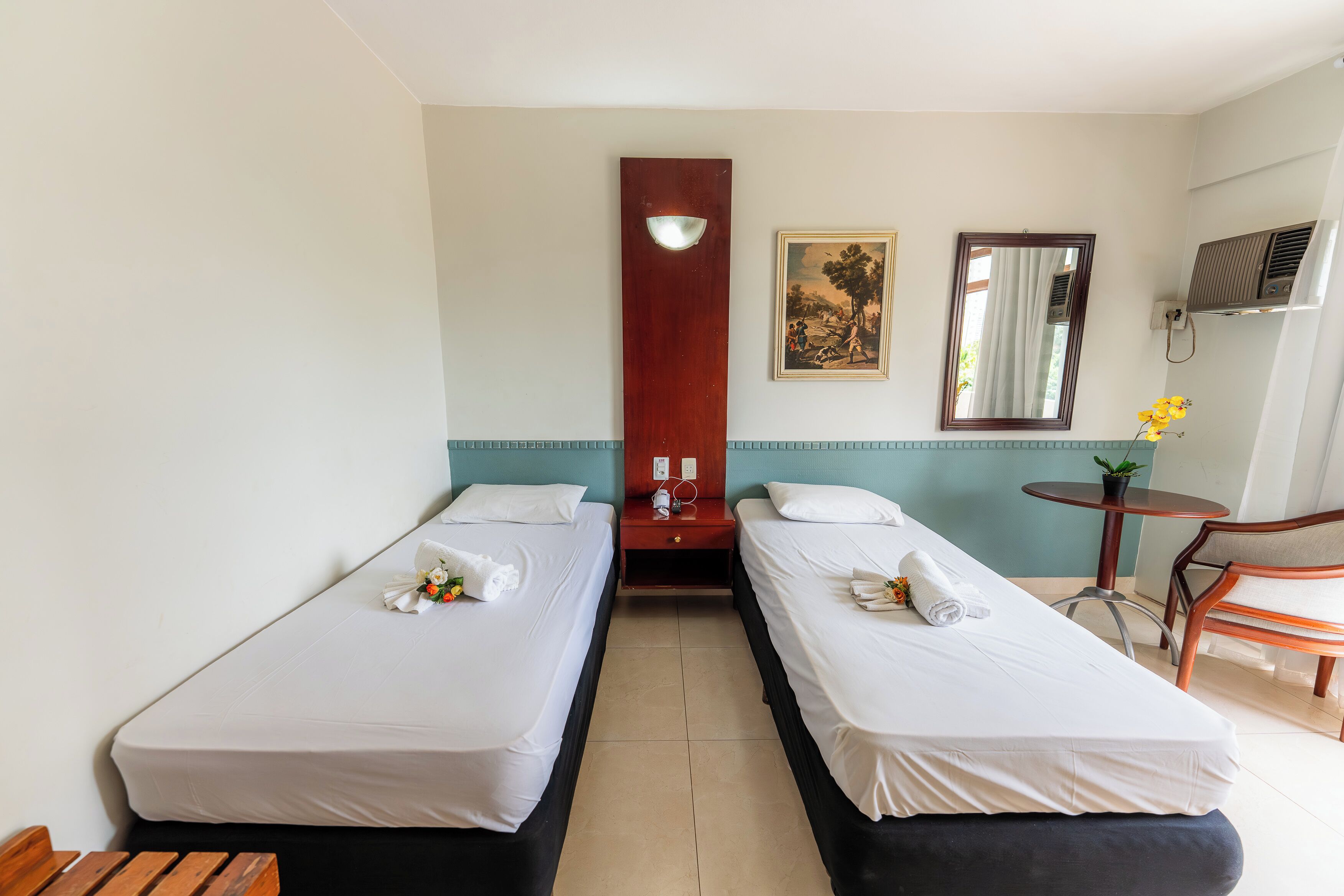 Een hotelkamer met twee eenpersoonsbedden, een houten nachtkastje, een klein tafeltje met een plant, een stoel en een wandgemonteerde airconditioning.
