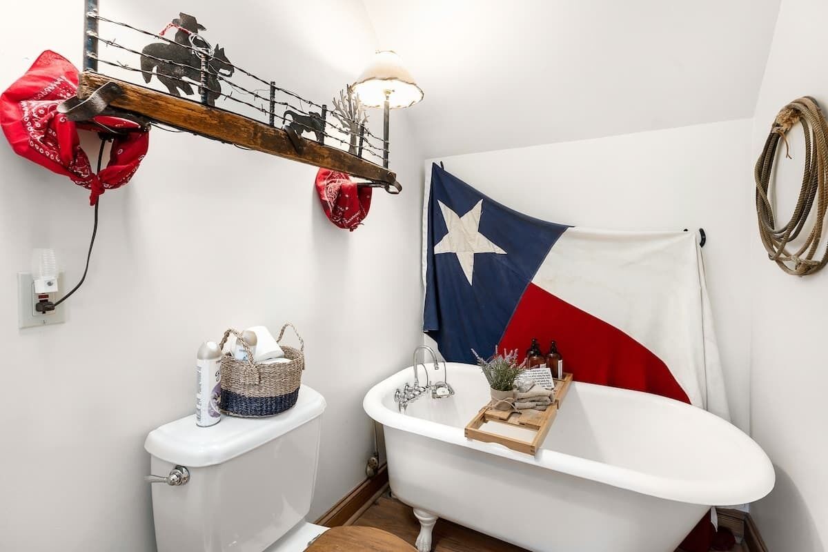 A bathroom with a white toilet, a vintage-style bathtub, a Texas flag, and a rustic shelf with a cowboy hat and a red bandana.