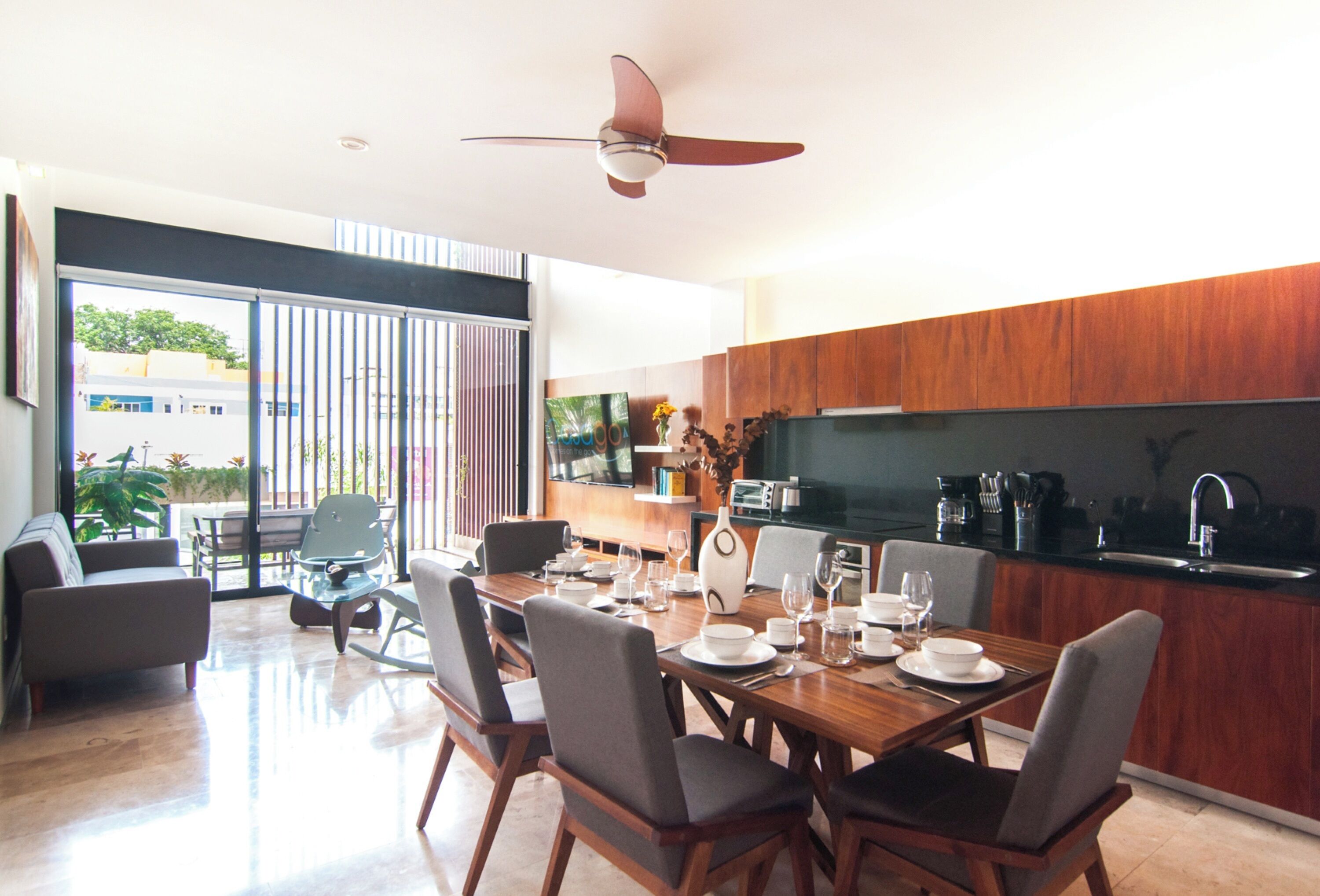 A modern kitchen with wooden cabinets, a dining area with a table set for four, and a large window providing a view of the outdoors.