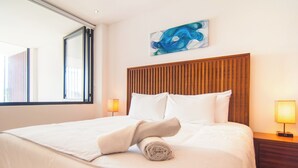 A neatly made bed with white linens and a rolled towel, a wooden headboard, and two bedside lamps on a nightstand. - Kuxtal 3 Bedroom Downtown Apartment  (Playa del Carmen)