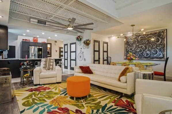 A modern living room with a white sofa, an orange ottoman, and a colorful rug. There is a kitchen in the background with a refrigerator and cabinets.
