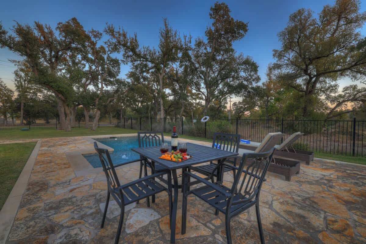 A stone patio with a pool, outdoor seating, and trees in the background.