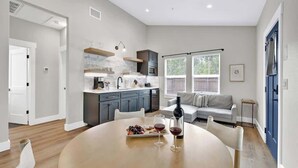 A modern kitchen with a dining area, featuring a round table with a wine bottle and glasses, and a sofa in the background.