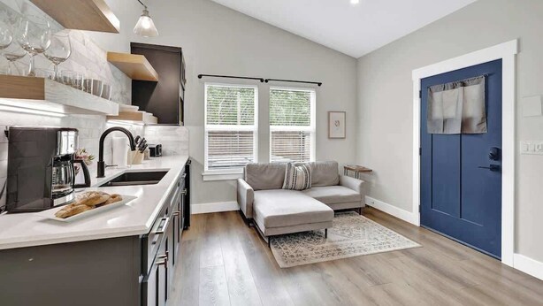 A modern kitchen with a built-in oven, a dining area with a white table and chairs, a grey sofa, and a blue door.