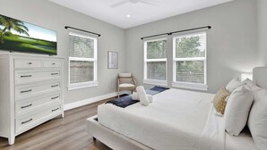 A bedroom with a bed, a TV, a dresser, and two windows with blinds.