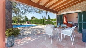 A covered outdoor patio with a pool, white wicker furniture, and a stone wall. - Villa Camarasa by Balearic Villas (Pollensa)