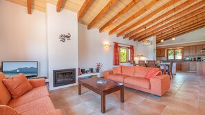 A spacious living room with a fireplace, a sofa, a coffee table, and a television. There is a kitchen in the background with wooden cabinets and a dining area. - Villa Camarasa by Balearic Villas (Pollensa)