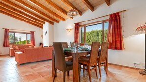 A dining area with a wooden table and chairs, a sofa, and a large window with curtains. - Villa Camarasa by Balearic Villas (Pollensa)