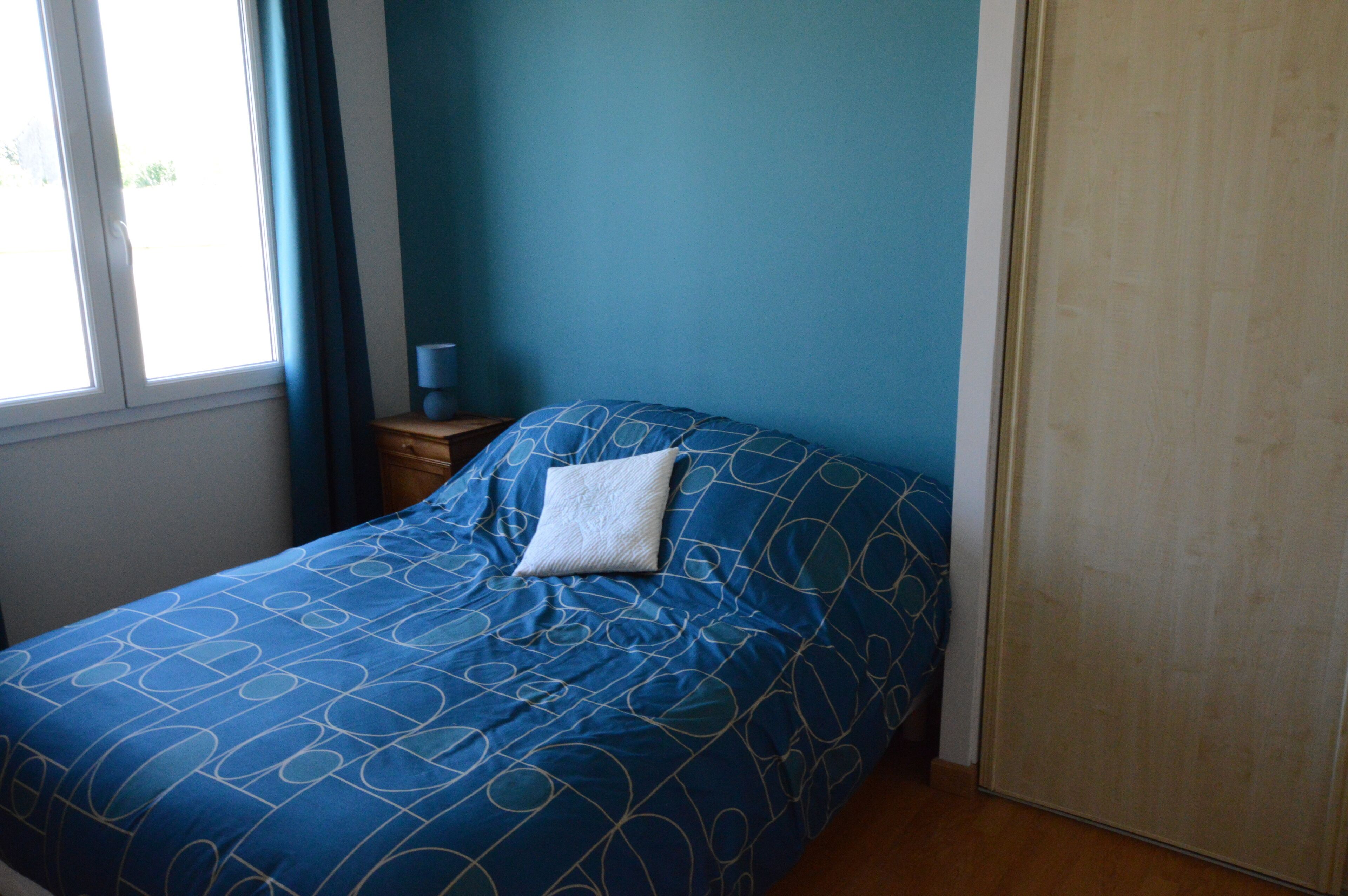 Un dormitorio con una colcha azul, una almohada blanca, una mesita de noche con una lámpara y una puerta de madera.