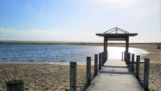 Ein hölzerner Steg führt zu einem Strand, am Ende befindet sich ein Holzbau.