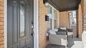 A front porch with a dark gray door, wicker chairs, and a brick wall.
