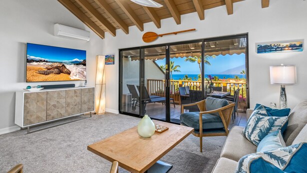 A living room with a TV, a wooden table, a sofa, and a view of the ocean through large glass doors. - Luxurious & Spacious Ocean Vistas (Kihei)