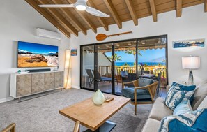 A living room with a TV, a wooden table, a sofa, and a view of the ocean through large glass doors. - Luxurious & Spacious Ocean Vistas (Kihei)