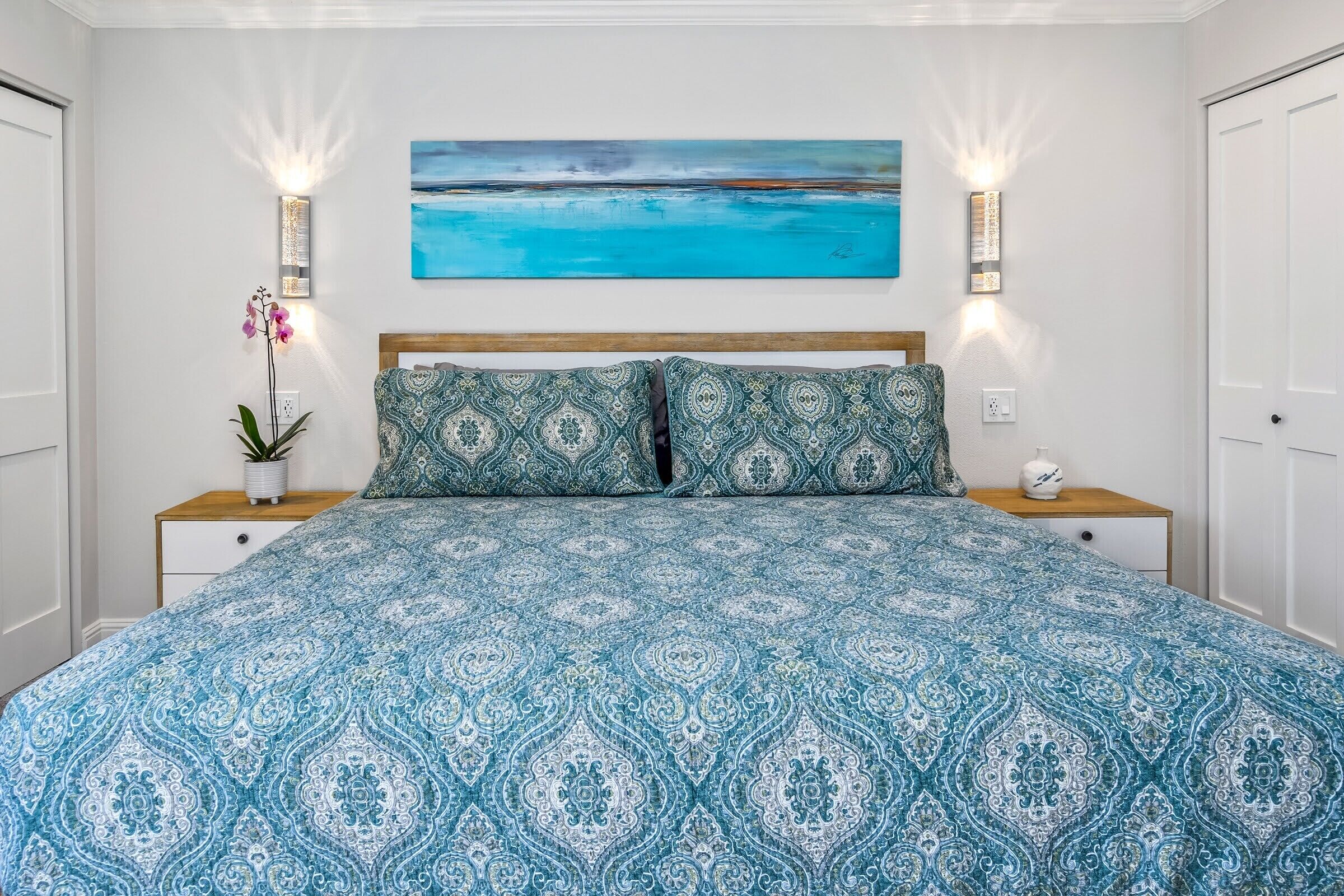 Un dormitorio con una cama grande, ropa de cama estampada, dos mesitas de noche con lámparas, una planta en maceta y un cuadro en la pared.