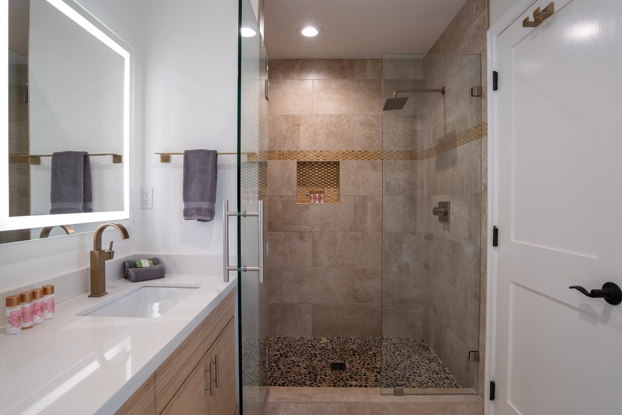 A modern bathroom with a glass shower enclosure, a large mirror, and a white sink with a gold faucet.