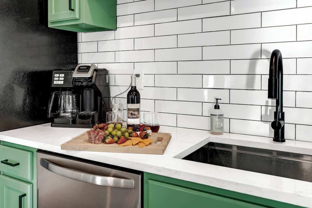 A kitchen with green cabinets, a white countertop, a coffee maker, a wine bottle, glasses, and a cutting board with fruits and cheese.